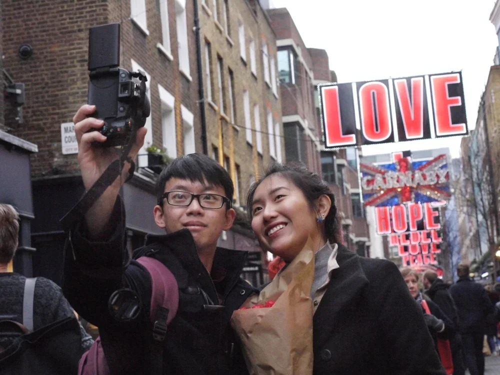 A young man and woman taking a selfie on a busy street with signs that read 'LOVE,' 'CANNABIS,' 'HOPE,' 'LOVE,' and 'PEACE' in the background.