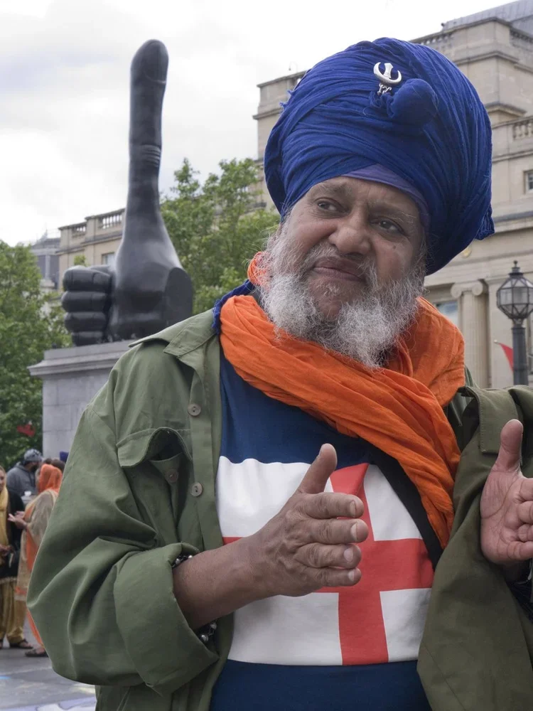 A man wearing a blue turban, orange scarf, and green jacket making a gesture with his hands, standing in front of a large black thumb sculpture.