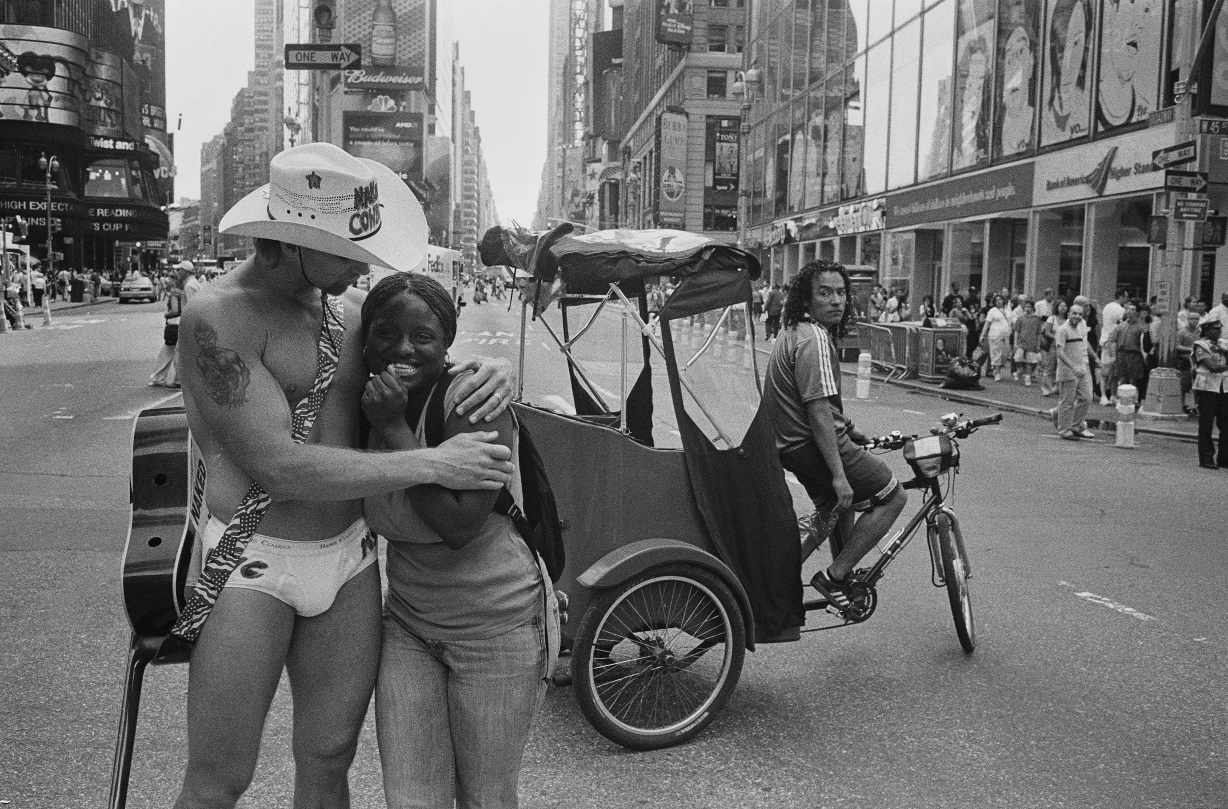 A black and white photo of two women hugging in the middle of a busy city street, with a young woman riding a bicycle nearby and a crowd of pedestrians in the background.