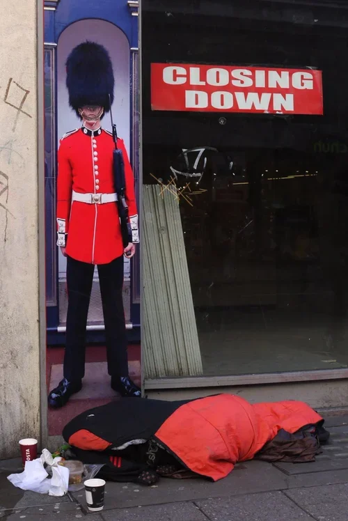 A person dressed as a British guard stands outside a store with a 'Closing Down' sign. The guard mannequin is in a red uniform with black hat and boots. In front, a person is sleeping on the sidewalk covered with a red and black blanket, with cups an