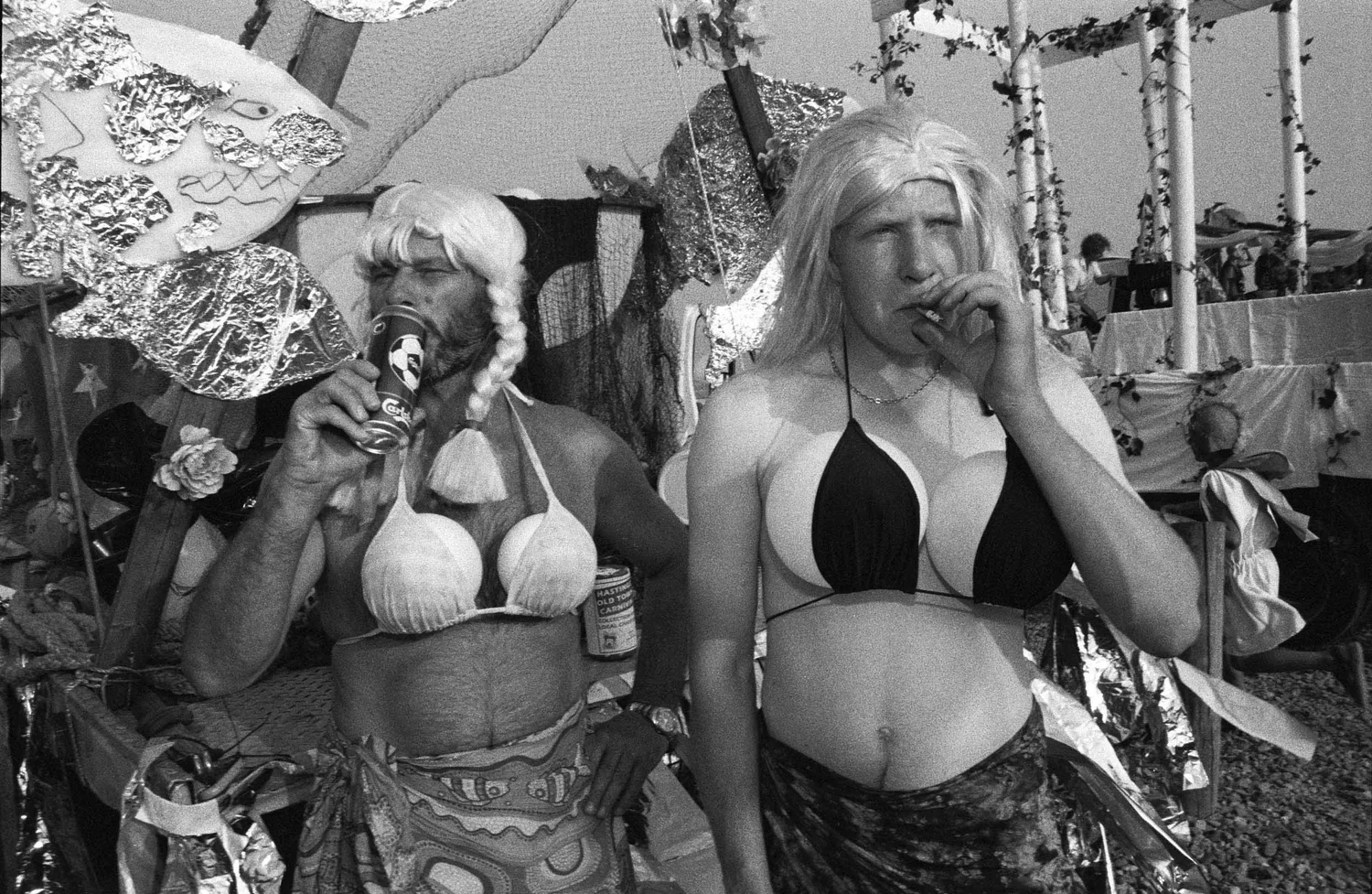 Two women in costumes—one dressed as a pirate with a wig and the other in a bikini top and skirt—at an outdoor event, with tents and other people in the background.