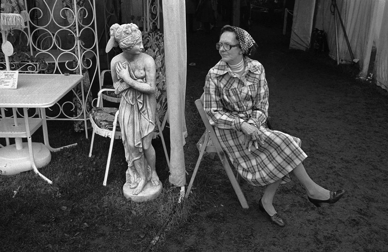 A woman sitting on a chair next to a classical statue of a woman against a decorative metal fence. The woman is wearing a plaid dress, glasses, and a headscarf, and is sitting with her legs crossed.
