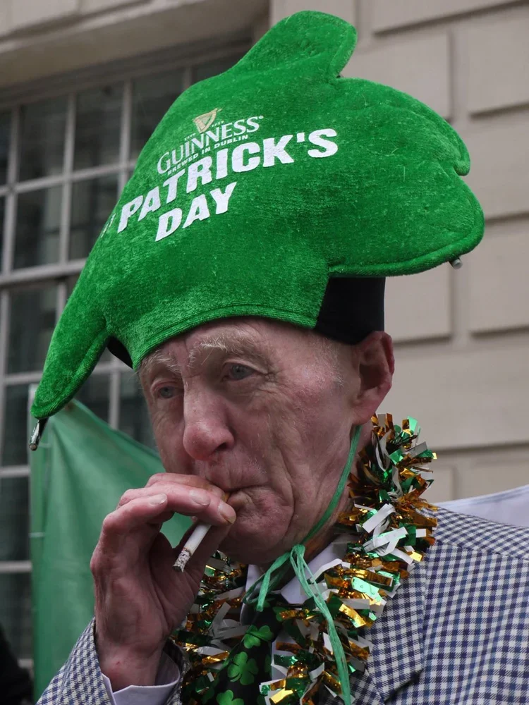 An elderly man wearing a green Guinness shamrock hat with the words "Patrick's Day" on it, a checkered jacket, and a green, gold, and white tinsel garland around his neck, smoking a cigarette.