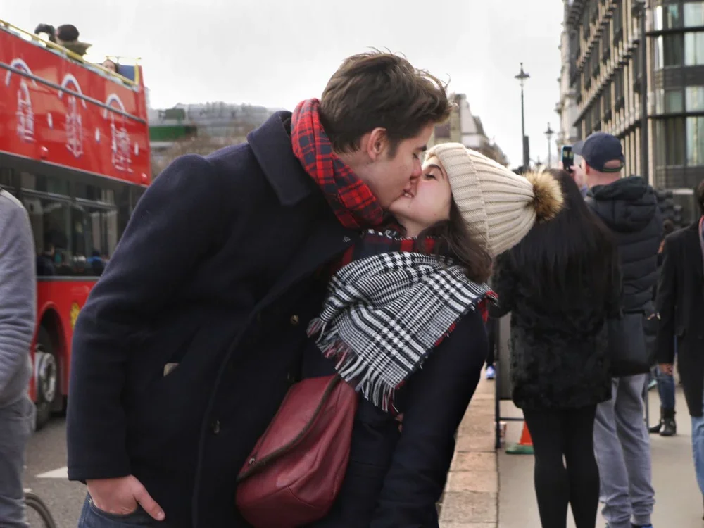 A young couple sharing a kiss on a busy city street during daytime, with a red double-decker bus and other pedestrians in the background.
