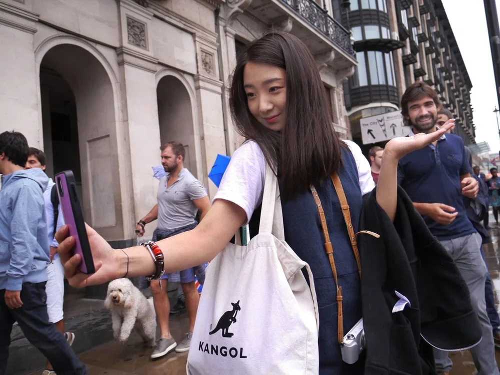A young woman taking a selfie on a busy city street, with several pedestrians and a small white dog in the background.