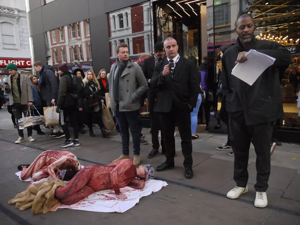 Two street performers dressed as homeless people lying on the ground, with a crowd of people standing nearby and observing, outside a building with glass windows.
