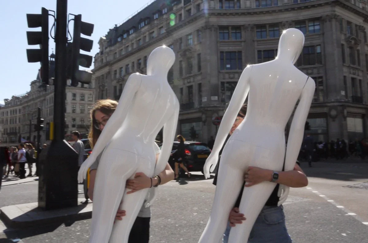 Two people holding large white mannequins in an urban street scene with tall buildings and traffic lights.