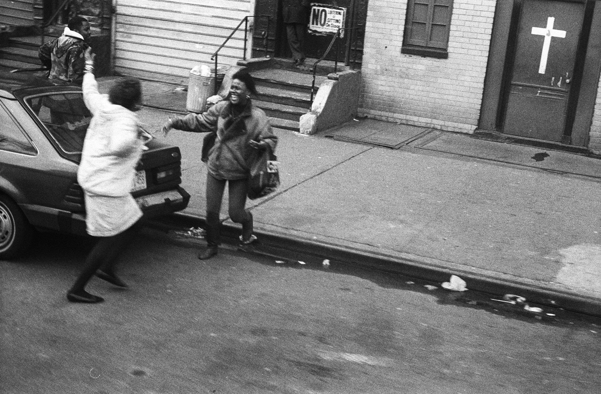 Two women on the sidewalk near a parked car are greeting each other with a handshake or high five. One woman appears to be smiling and holding a purse. The scene takes place outside a building with stairs, a sign that says "No," and a religious cross