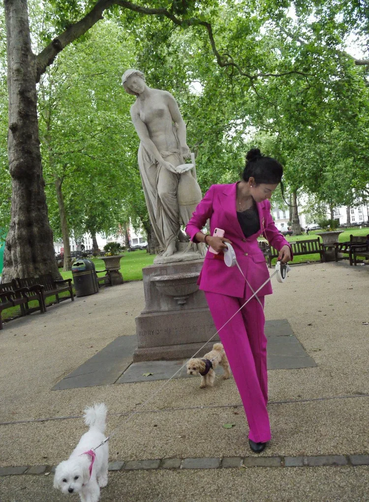 Woman in a bright pink suit walking two small dogs on leashes in a park with a classical statue of a woman pouring water from a jar behind her. Trees and benches are visible in the background.