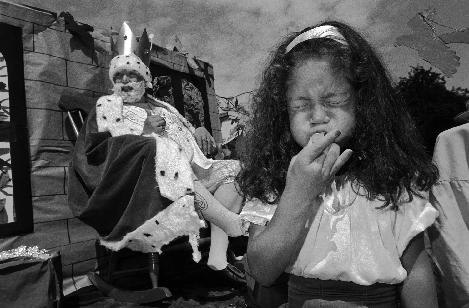 A young girl with curly hair and a headband, squinting with eyes closed, eating a snack outdoors during daytime. Behind her, a person dressed as a king, wearing a crown and royal robes, is seated in front of a decorated backdrop.