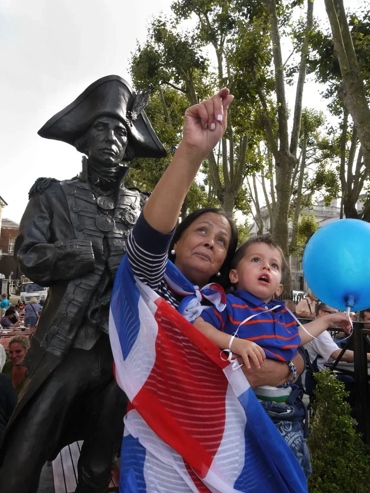 A woman and a boy in front of a statue of a historical military figure, both wrapped in a French flag. The woman is pointing upward, and the boy is holding a blue balloon.