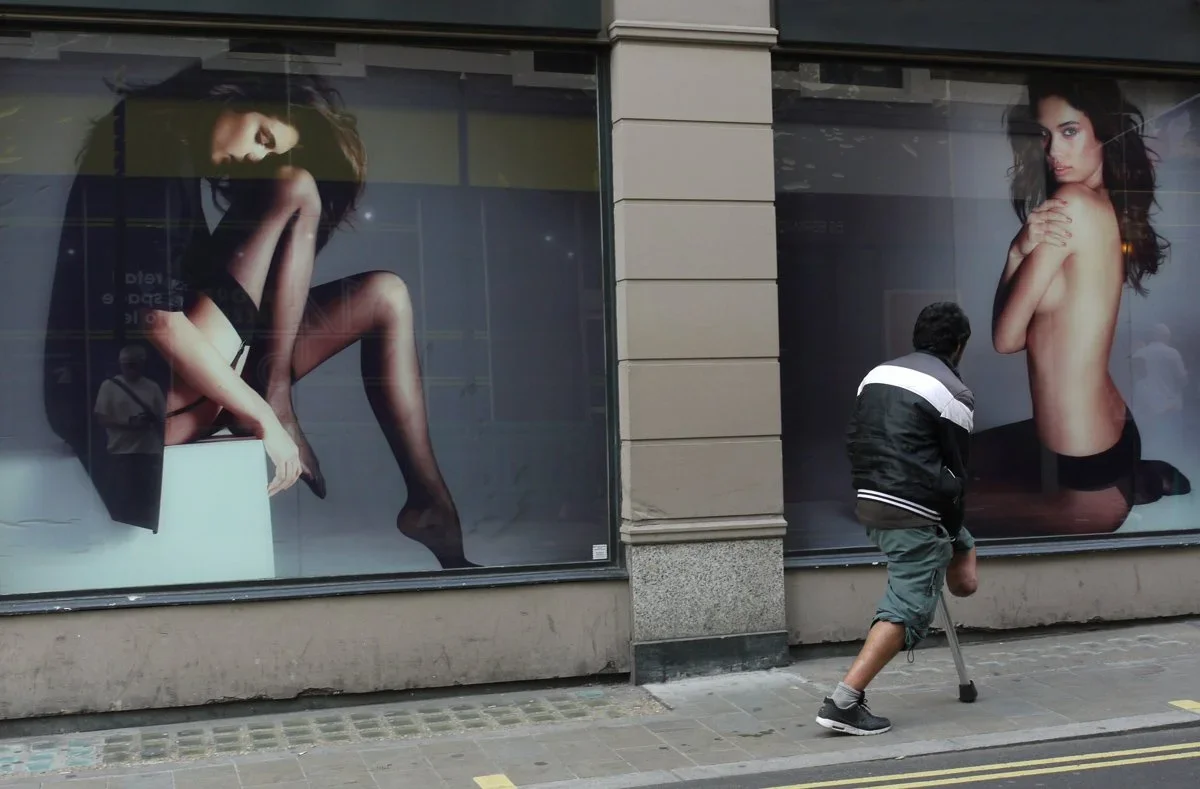 A person with a crutch walking past a storefront with large fashion advertisements featuring women in lingerie.