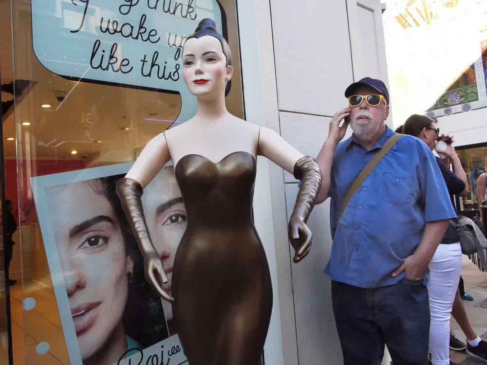 A man with sunglasses and a blue shirt talking on his cellphone stands next to a mannequin dressed in a metallic, strapless, form-fitting gown with long gloves, outside a store with posters and advertisements.