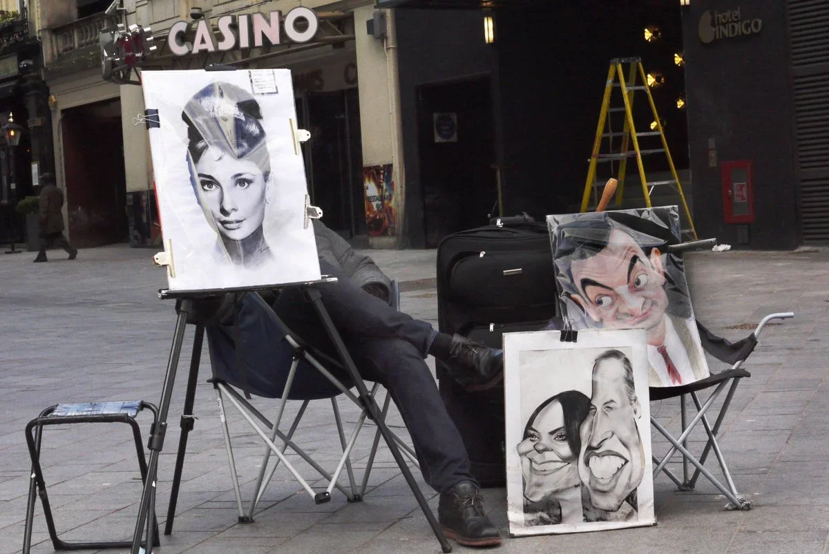 Street artist with caricature portraits on display, including a black-and-white portrait of a woman and exaggerated cartoon-style faces of a man and woman.