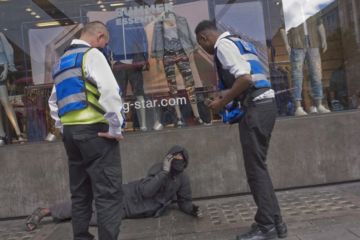 A person in black clothing and a face mask lies on the sidewalk, appearing to be homeless, while two security officers stand nearby and a man records the scene with a phone in front of a clothing store window.
