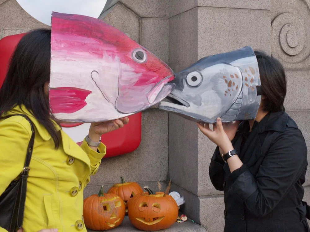Two women are holding large fish-shaped masks in front of their faces, making it appear as if they are kissing fish. The women are standing outside near a wall with carved designs, and there are three painted pumpkins on the ground below.