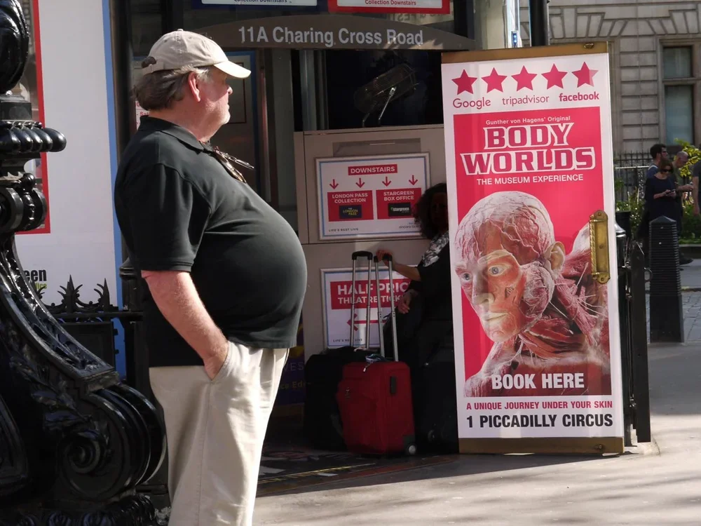 A man with a gray cap, black shirt, and beige pants standing outside a ticket booth for Body Worlds exhibit at Piccadilly Circus, London, with a large promotional sign showing a human body with muscles and organs.