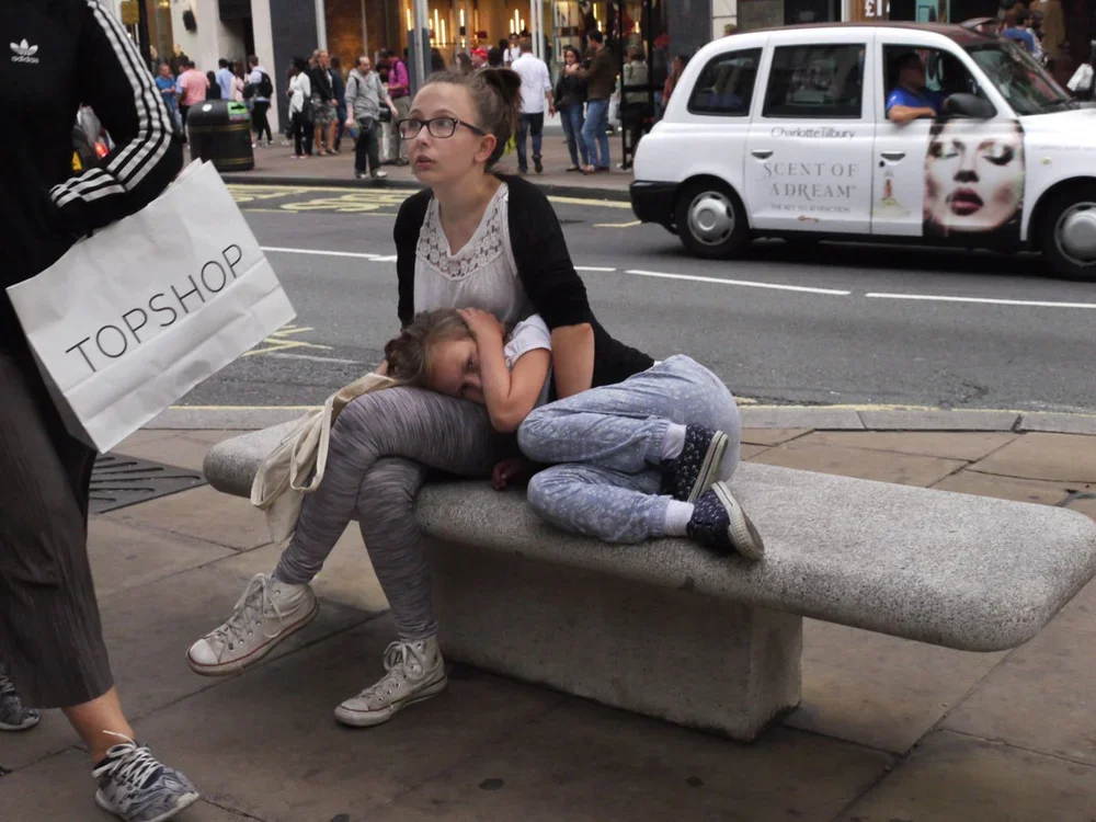 Two young girls sit on a stone bench on a city sidewalk, with one girl resting her head on the other's lap. The girl sitting up has glasses and wears a white top and dark cardigan. The girl lying down appears to be sleeping or resting, wearing gray l