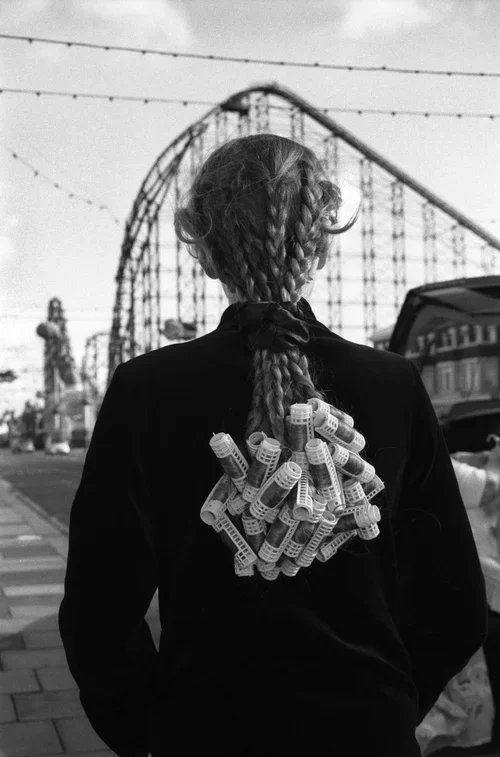 A person with braided hair carries hair rollers on their back, standing in front of a roller coaster at an amusement park.