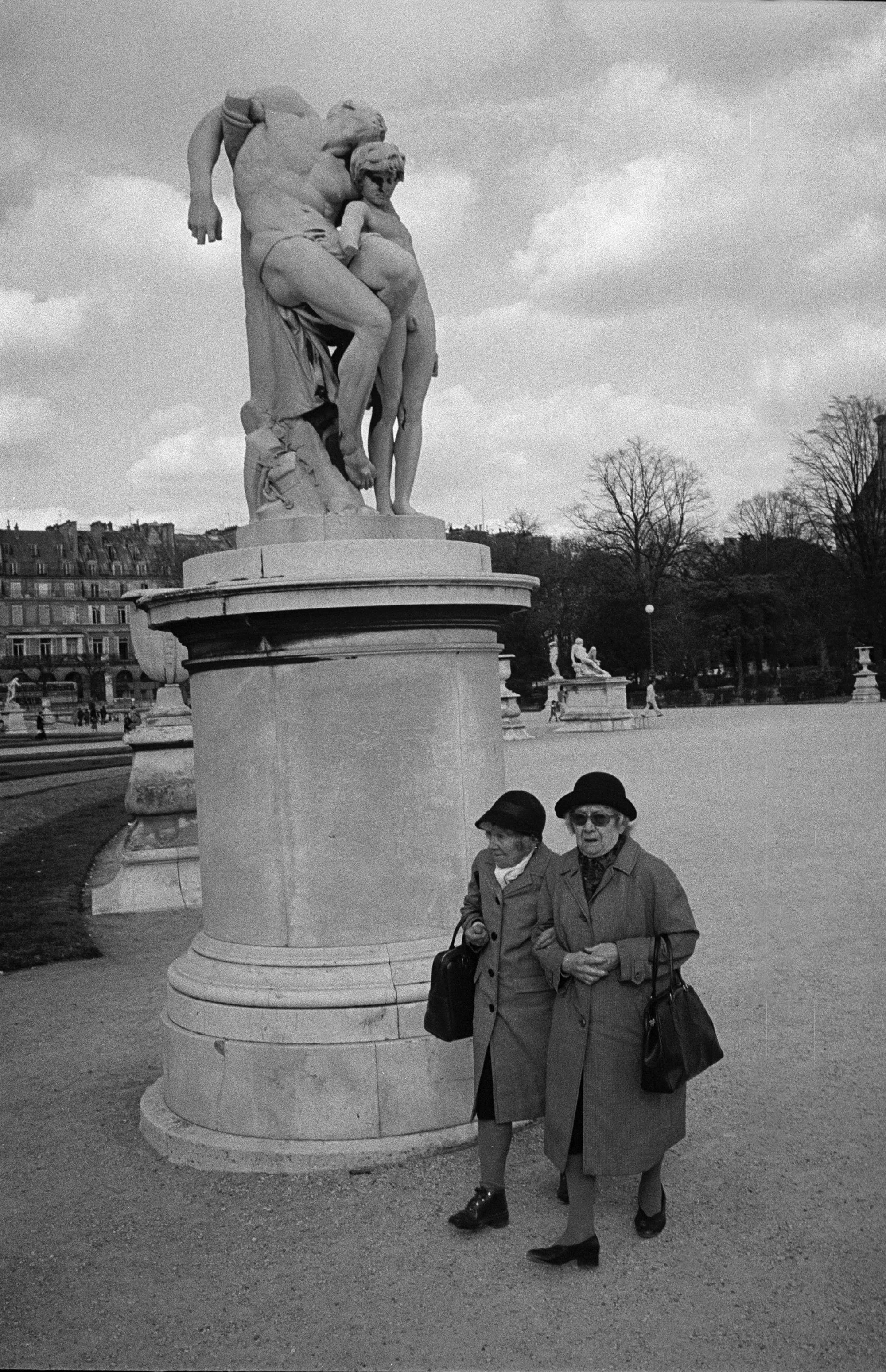 Black and white photo of two elderly women walking past a large classical statue with two children, situated on a pedestal in a park. In the background, there are trees and buildings, and a cloudy sky.