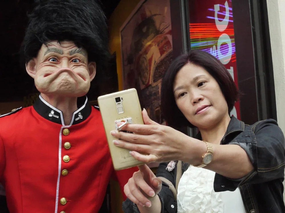 A woman taking a selfie with a man dressed in a costume of a police officer in a red uniform with gold buttons and a large distorted mask face in an outdoor or semi-outdoor setting.