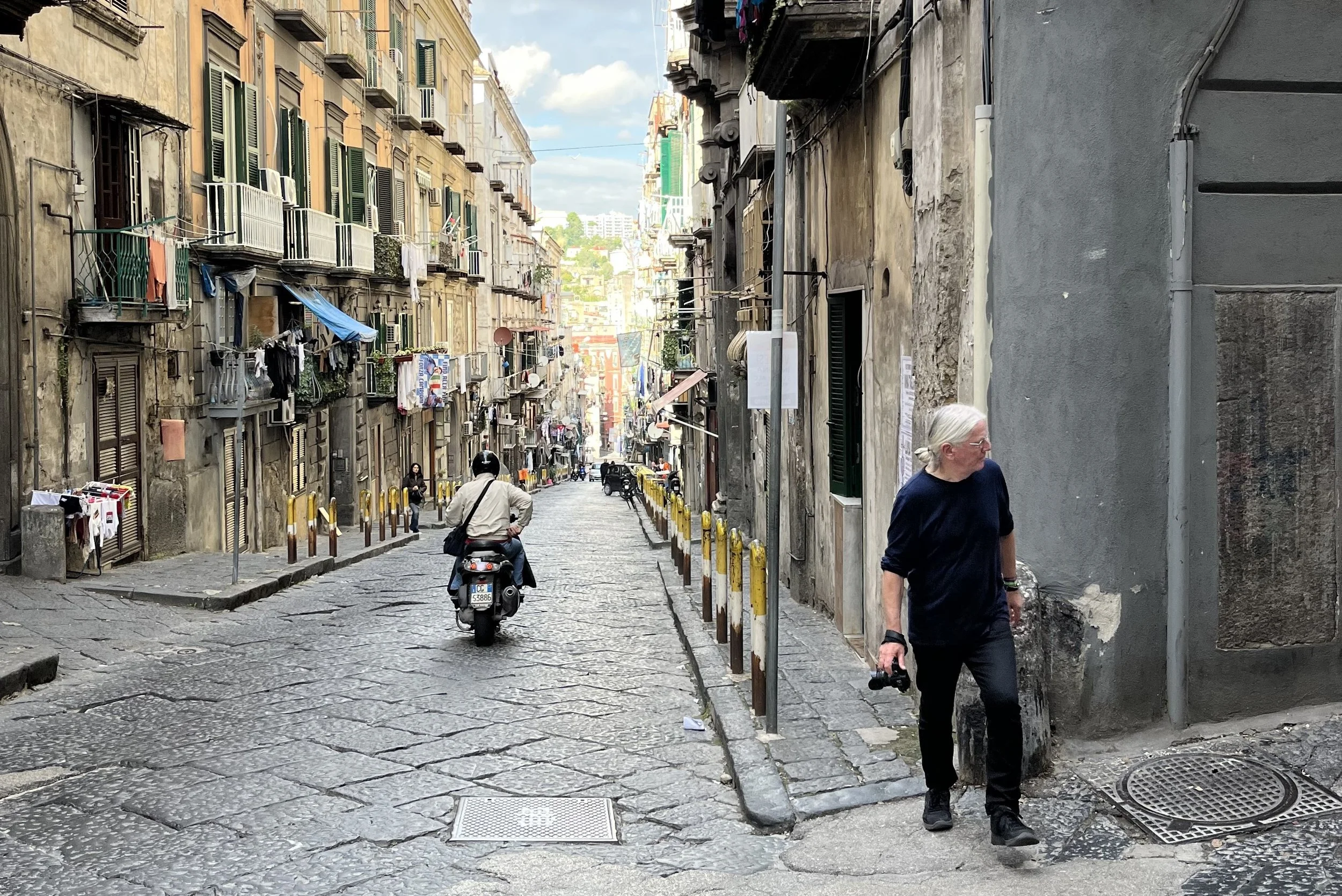 A narrow cobblestone street in an old urban neighborhood with colorful, aged buildings on either side. People are walking and riding a scooter, and laundry hangs from balconies.