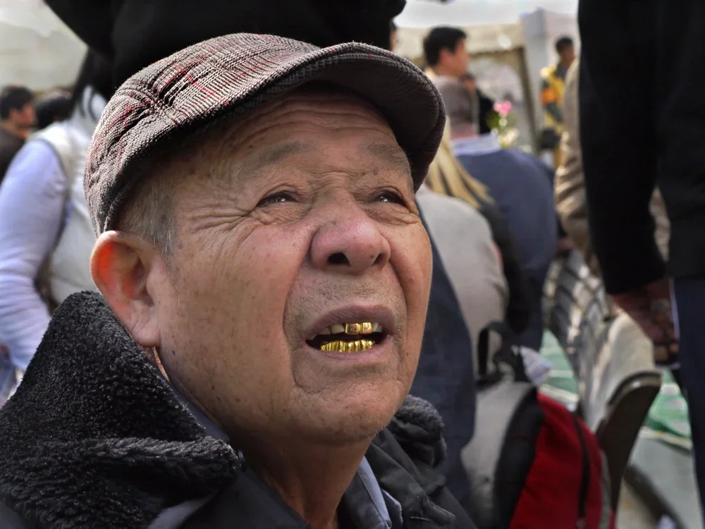 An elderly man with gold teeth, wearing a plaid cap and a dark fuzzy coat, looking upwards in a crowded outdoor setting.
