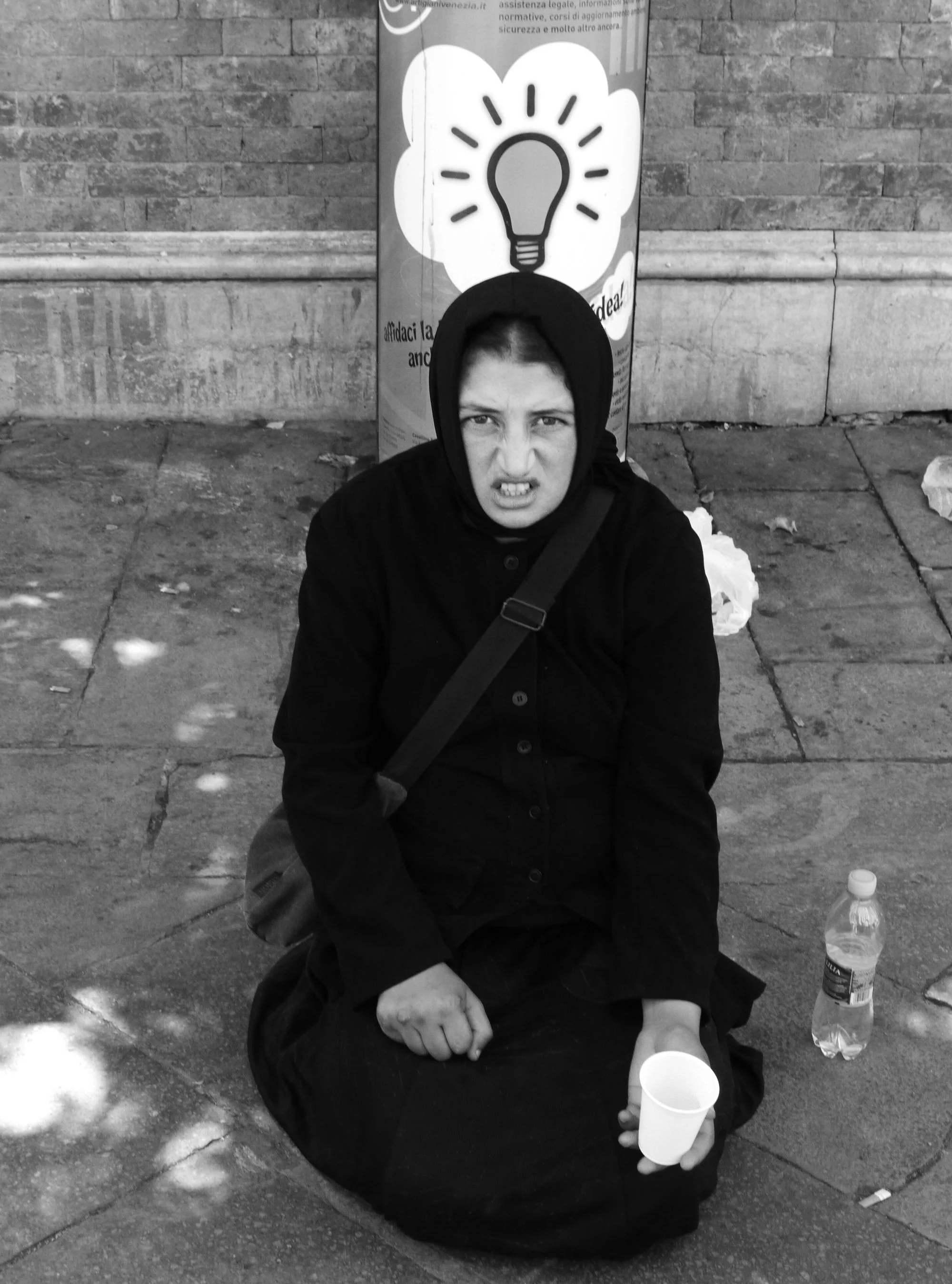 A person in a black hoodie and coat sitting on the sidewalk, making a grimace while holding a paper cup, with a water bottle and trash visible on the ground; a brick wall and a poster with a lightbulb icon are in the background.