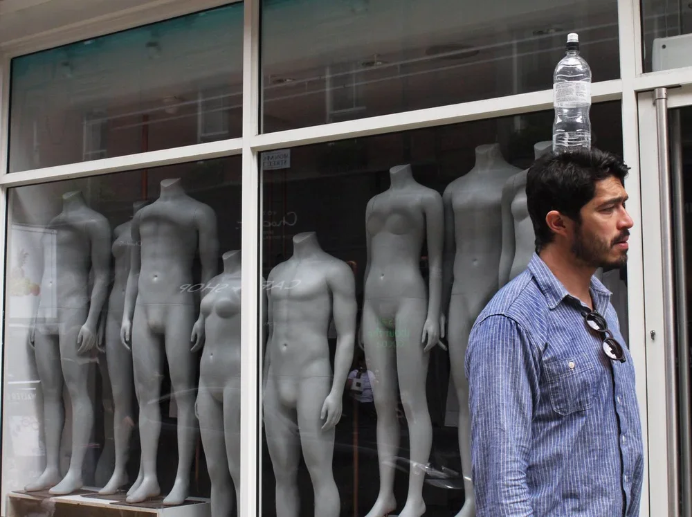 Man with sunglasses hanging from shirt collar walking past mannequin shop window with six blank female mannequins.