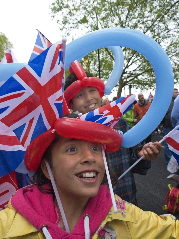 Children participating in a celebration, wearing National Day hats, balloons, and waving British flags during an outdoor event.