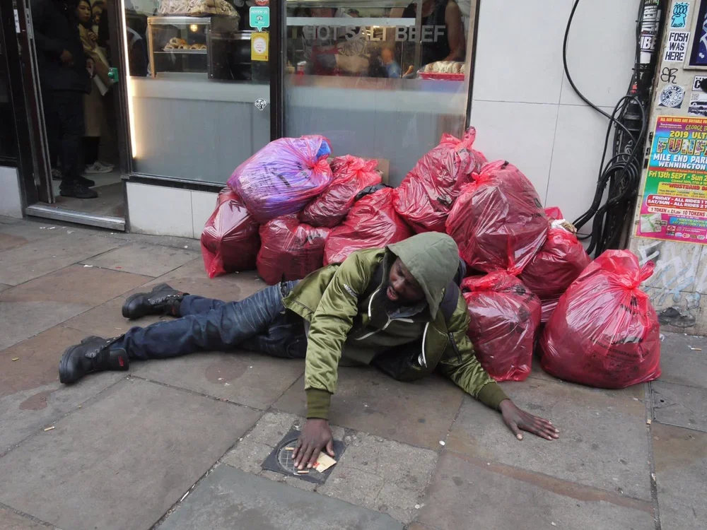 A man lying on the sidewalk next to a pile of red plastic bags filled with items outside a store.