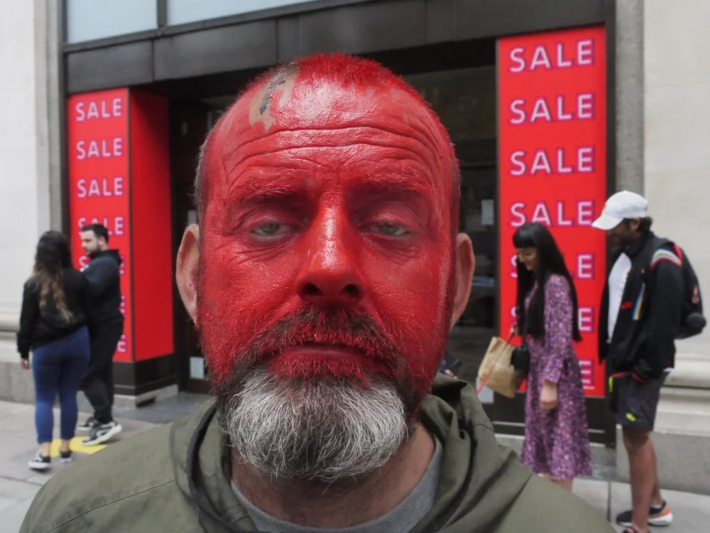 Man with his face painted entirely red standing in front of a store with sale signs and people walking by.