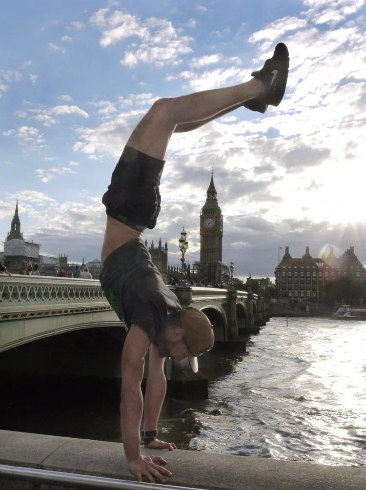 A man performs a handstand on the ledge of a bridge over the River Thames in London, with Big Ben and the Houses of Parliament in the background.