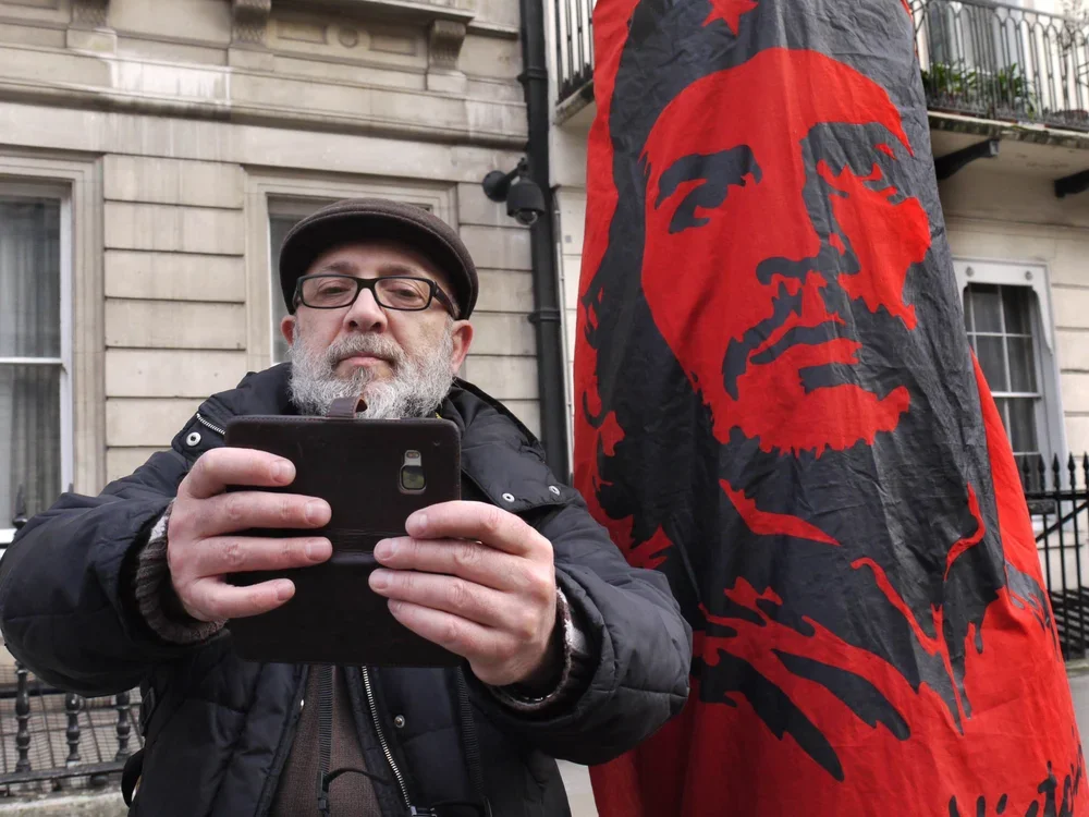 A man with glasses and a beard taking a selfie with a smartphone, standing next to a large red and black rally flag or banner with a portrait of a woman and stars.