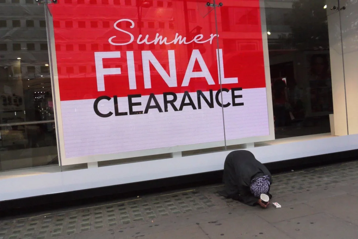 A person crouched on the sidewalk in front of a store window, near a large red and white sign that reads "Summer Final Clearance."