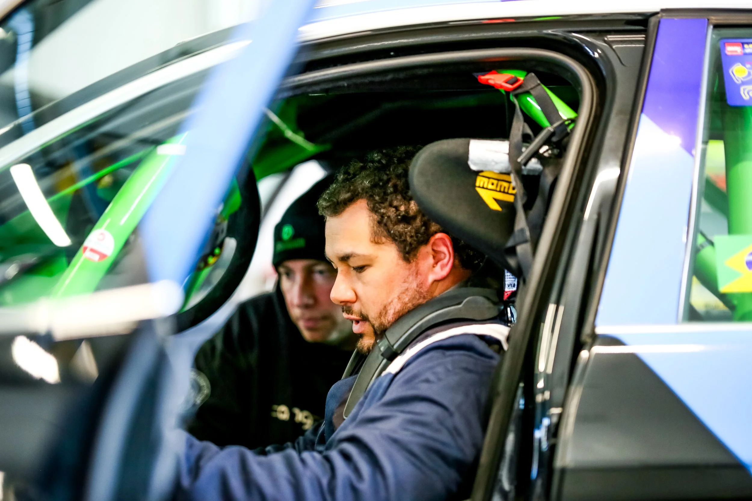 Close-up of two race car drivers sitting in their cars, wearing helmets. The driver in the foreground has a focused expression and curly hair visible under his helmet, while the other driver appears behind him.