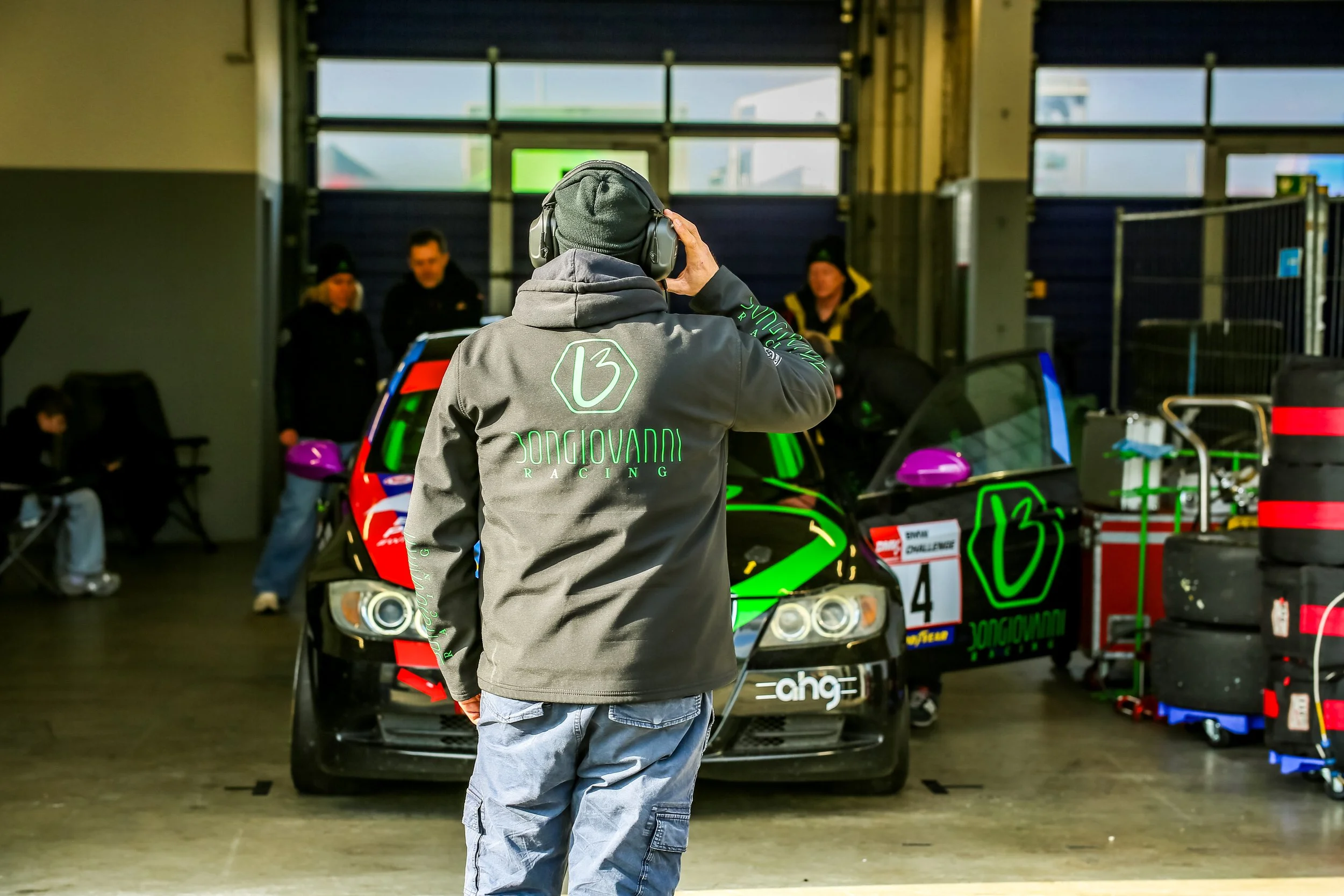 A man wearing a gray hoodie with green text and a gray beanie is speaking into headphones in a garage or workshop. In front of him are two race cars with green accents, and behind him are two people and various equipment.