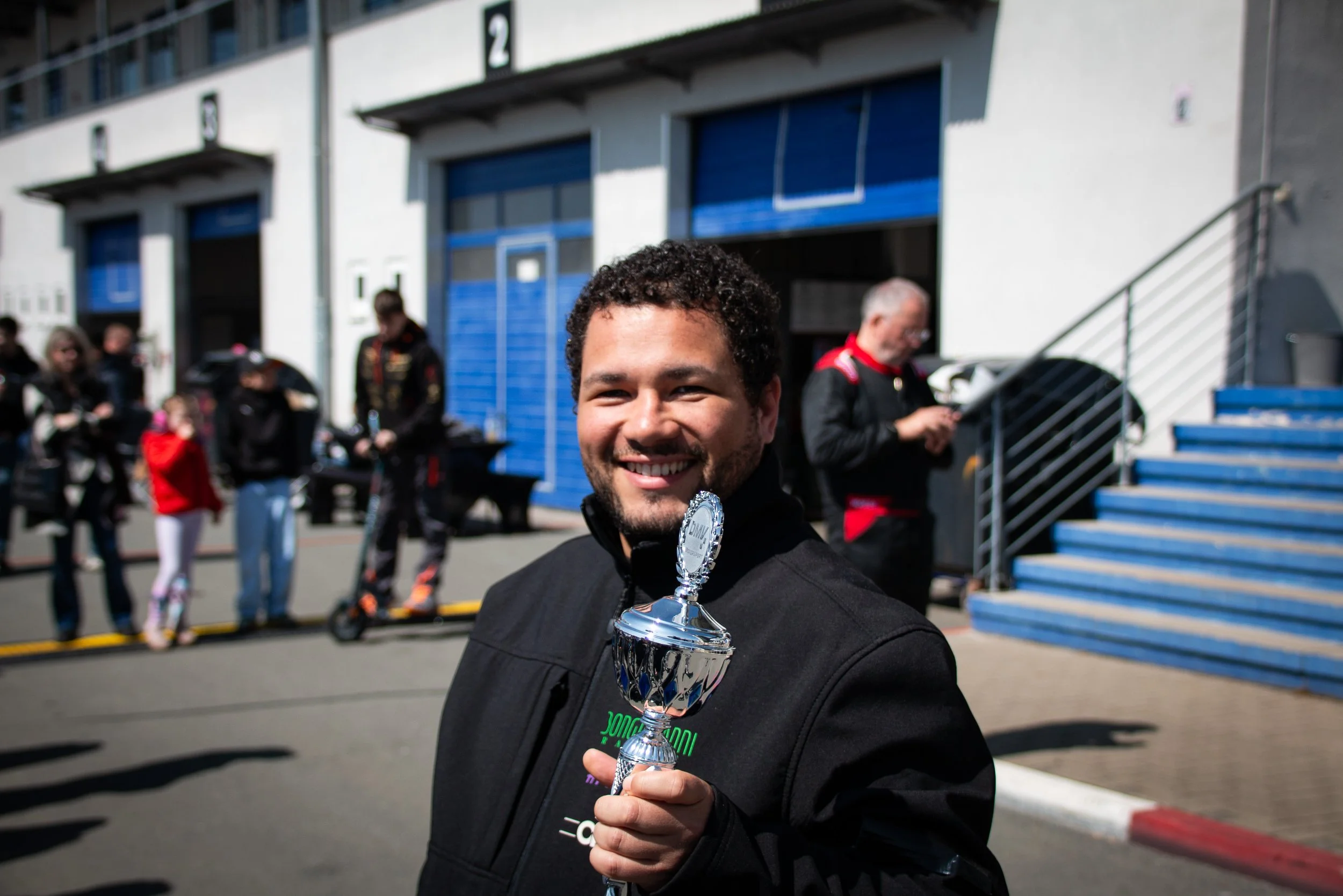 A smiling man holding a silver trophy at a race track with other people and race cars in the background.