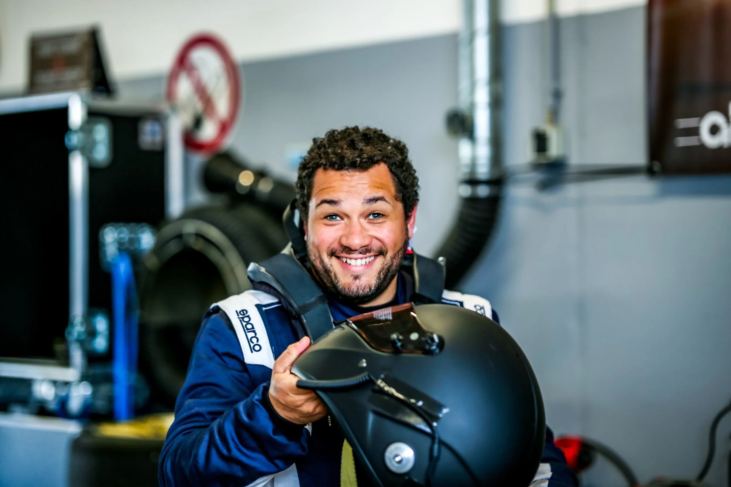 A smiling man with curly hair and a beard in a racing suit holding a black motorcycle helmet in a garage or pit lane.