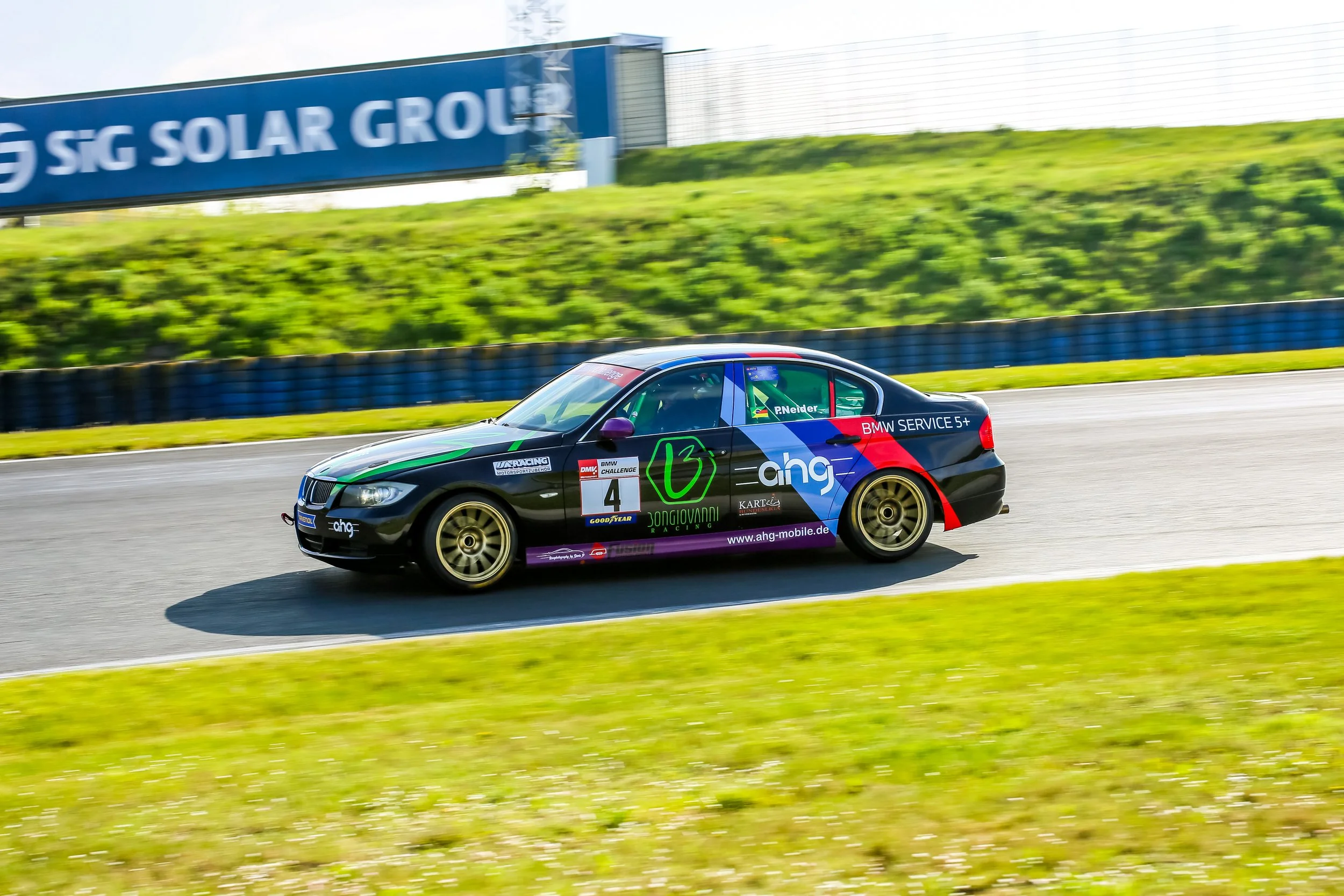 A race car on a racing track during a speed event, with a large blue sign in the background displaying 'SG SOLAR GROUP' and a grassy hill behind it.