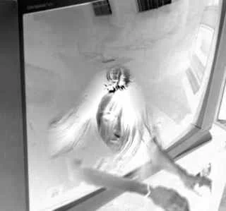A person with light-colored hair looking up at a camera through a glass door or window.