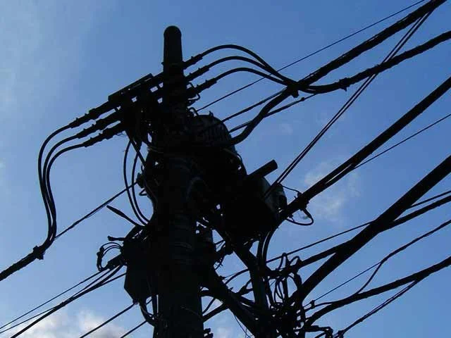 Silhouette of an electric utility pole with multiple power lines against a blue sky.