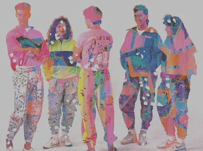 Group of six young adults standing together, wearing colorful casual clothing, against a plain white background.