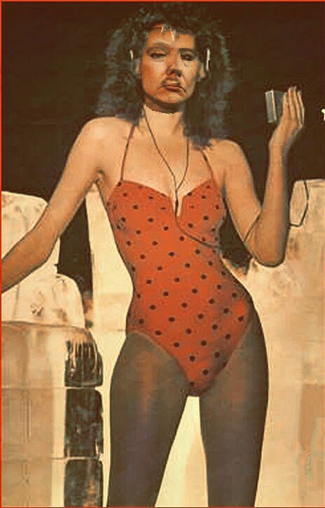 A woman with curly hair, wearing headphones, a red polka dot bodysuit, and black tights, posing with one arm resting on a stone pillar and the other holding a small object, in front of a brick wall.