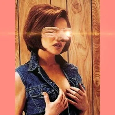 A woman with short brown hair taking a mirror selfie in front of a wooden wall, wearing a sleeveless denim jacket, with a bright flash obscuring part of her face and chest.