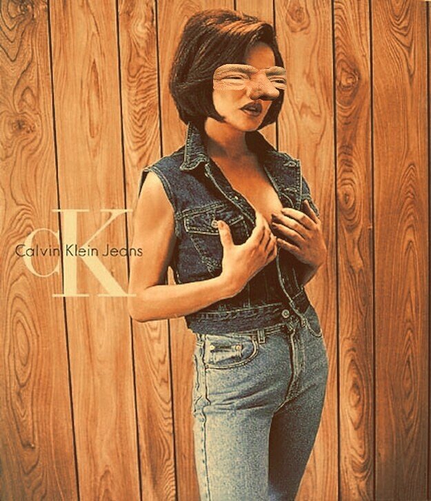 A woman with short dark hair and distorted facial features due to a filter, wearing a denim sleeveless vest and high-waisted jeans, standing against a wooden paneled wall, holding her chest. The text 