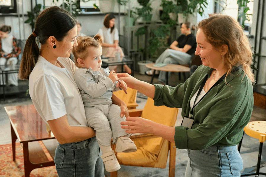 A woman holding a young toddler, both facing a smiling woman who is reaching out to the child in a social setting with other people in the background.
