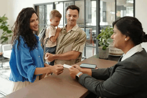 A woman at a reception desk handing a card to a smiling couple with a young child. The woman at the desk is wearing a gray blazer, and the couple is casually dressed. The setting appears to be a modern office or waiting area with large windows, plants, and a smartphone on the desk.