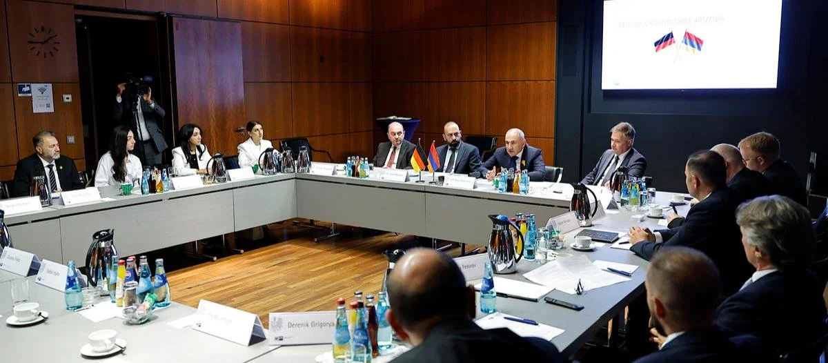 Menschen sitzen an einem großen Konferenztisch bei einer offiziellen Besprechung oder einem Treffen, einige mit kleinen Flaggen vor sich, in einem modernen Raum mit Holzpaneelen und einer großen Bildschirmanzeige im Hintergrund.