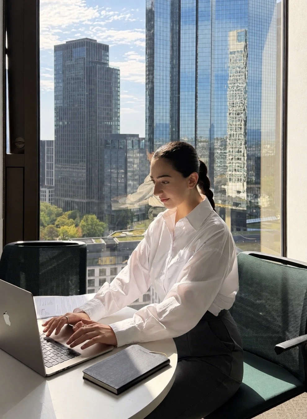 Frau arbeitet an einem Laptop in einem modernen Büro mit Blick auf hohe Glasgebäude im Stadtzentrum bei Sonnenschein.